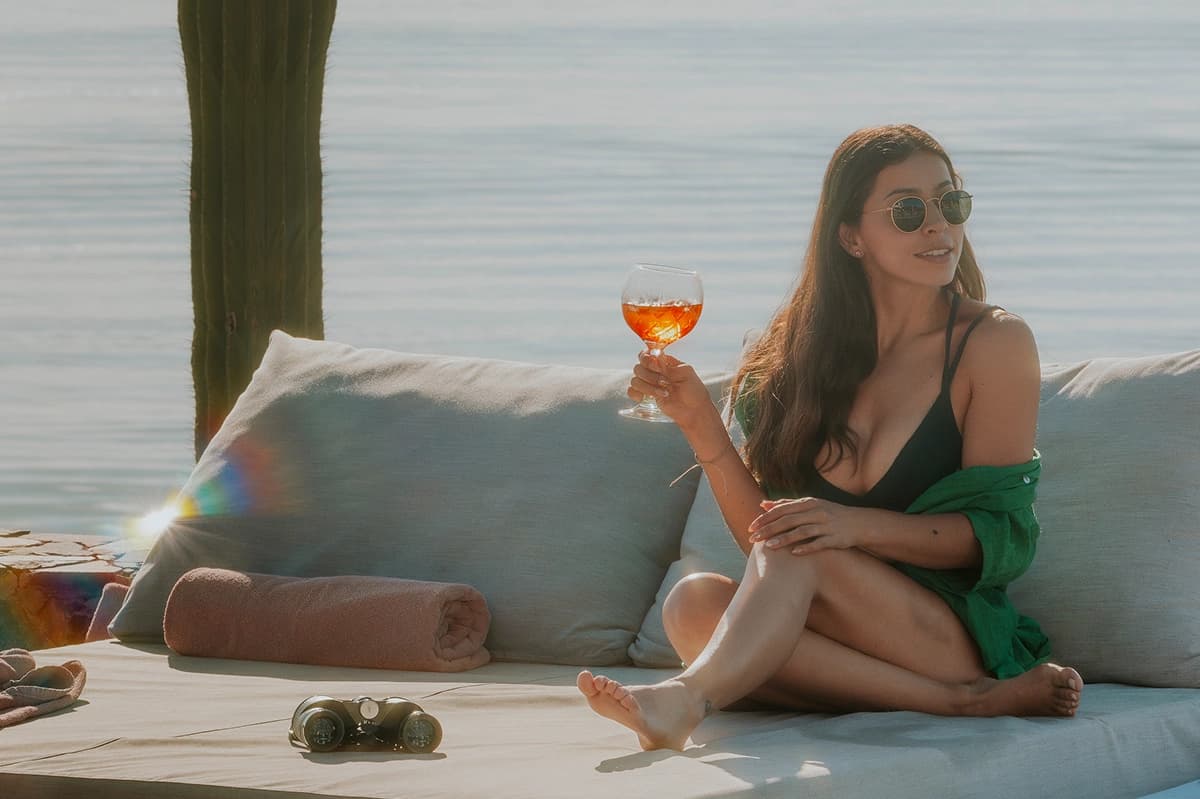 Alguien con lentes de sol sentado en un camastro frente al mar, con una copa de aperitivo y unos binoculares al lado
