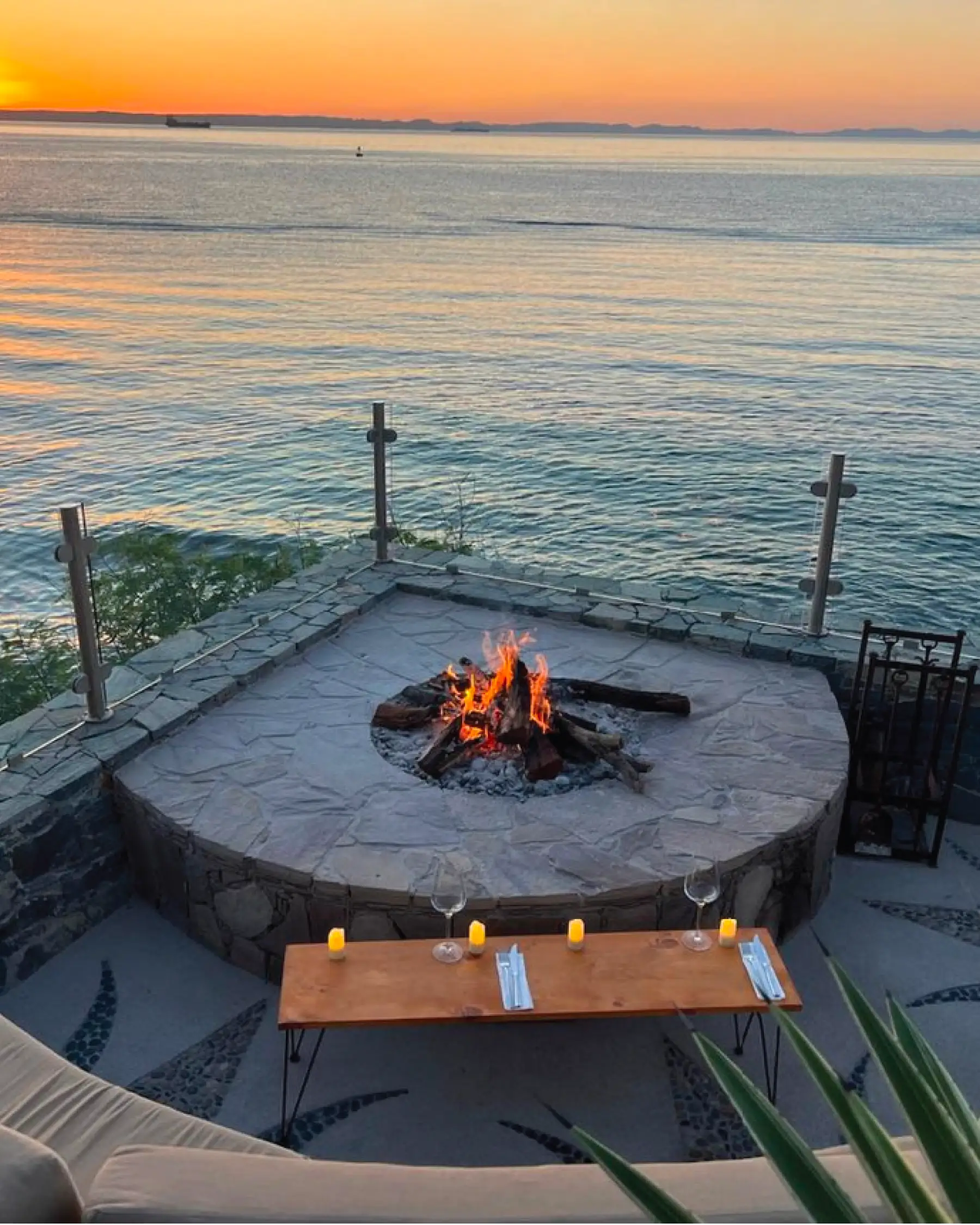 Stone terrace above the water with a lit fire pit, a low table with glasses and candles, and the sun setting over the bay