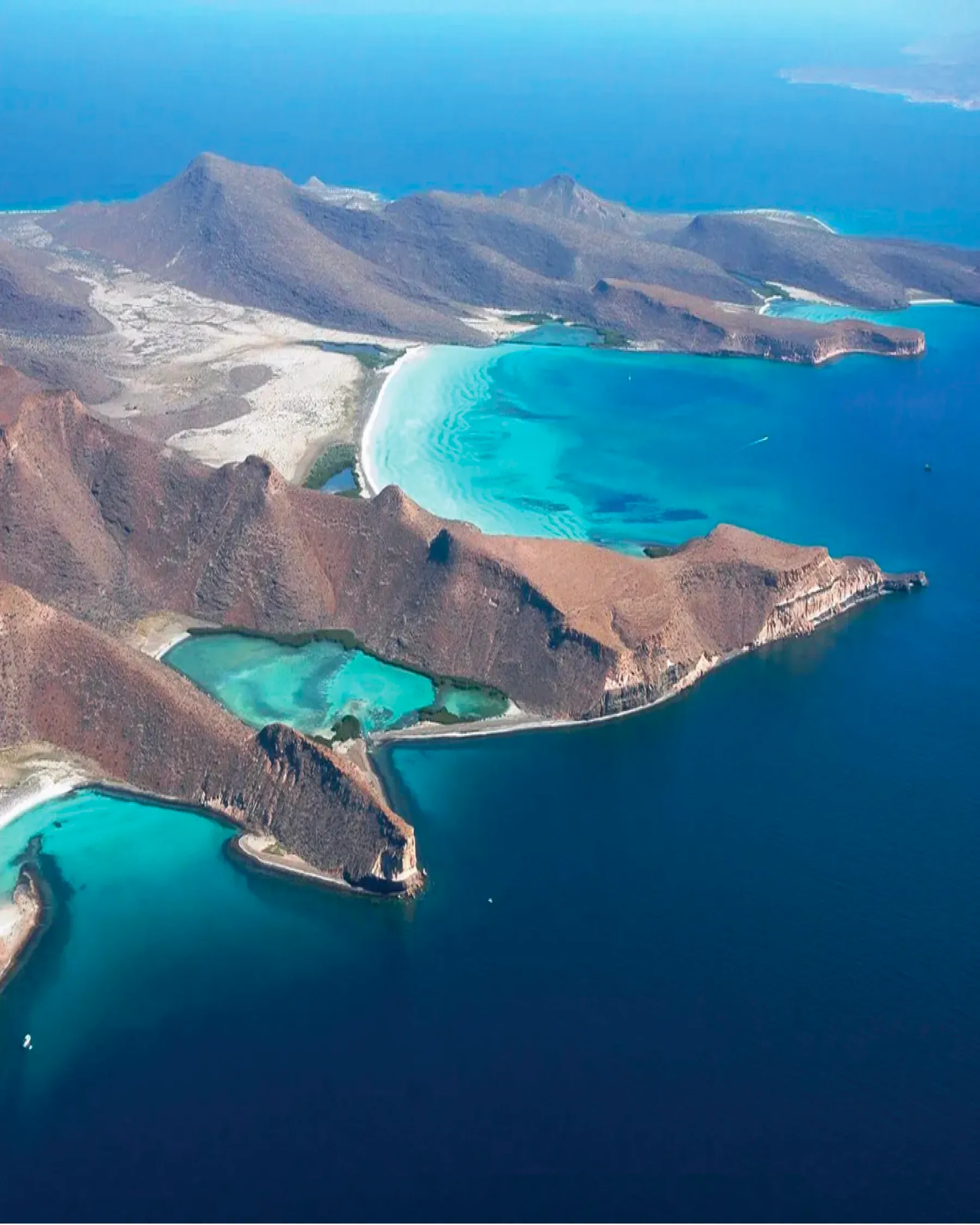 Aerial view of the island: brown rocky ridges dropping to the sea, with turquoise coves and deep blue water around them
