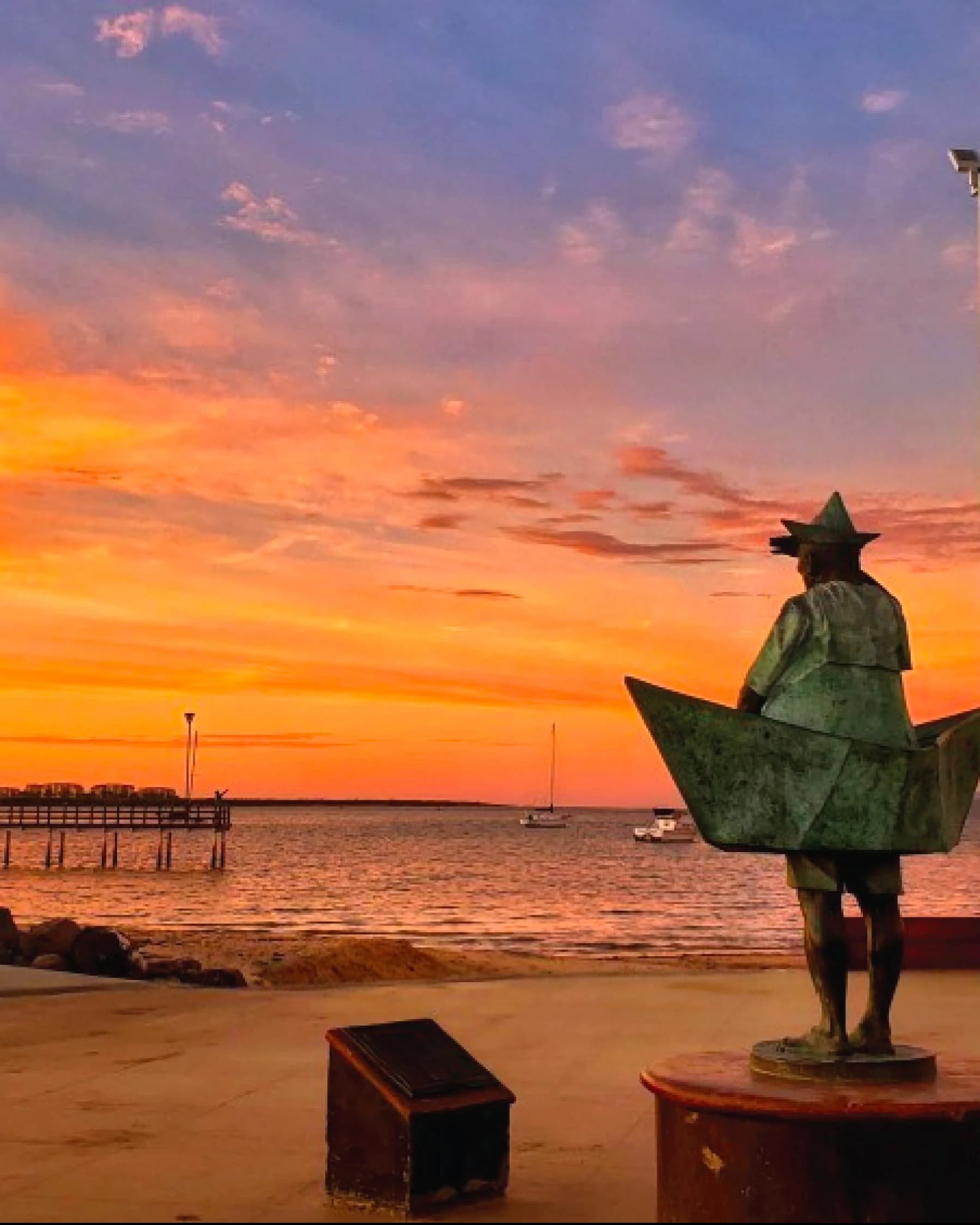 The La Paz malecón at sunset: the bronze sculpture of a figure inside a paper boat, with the pier and a few sailboats on the orange water
