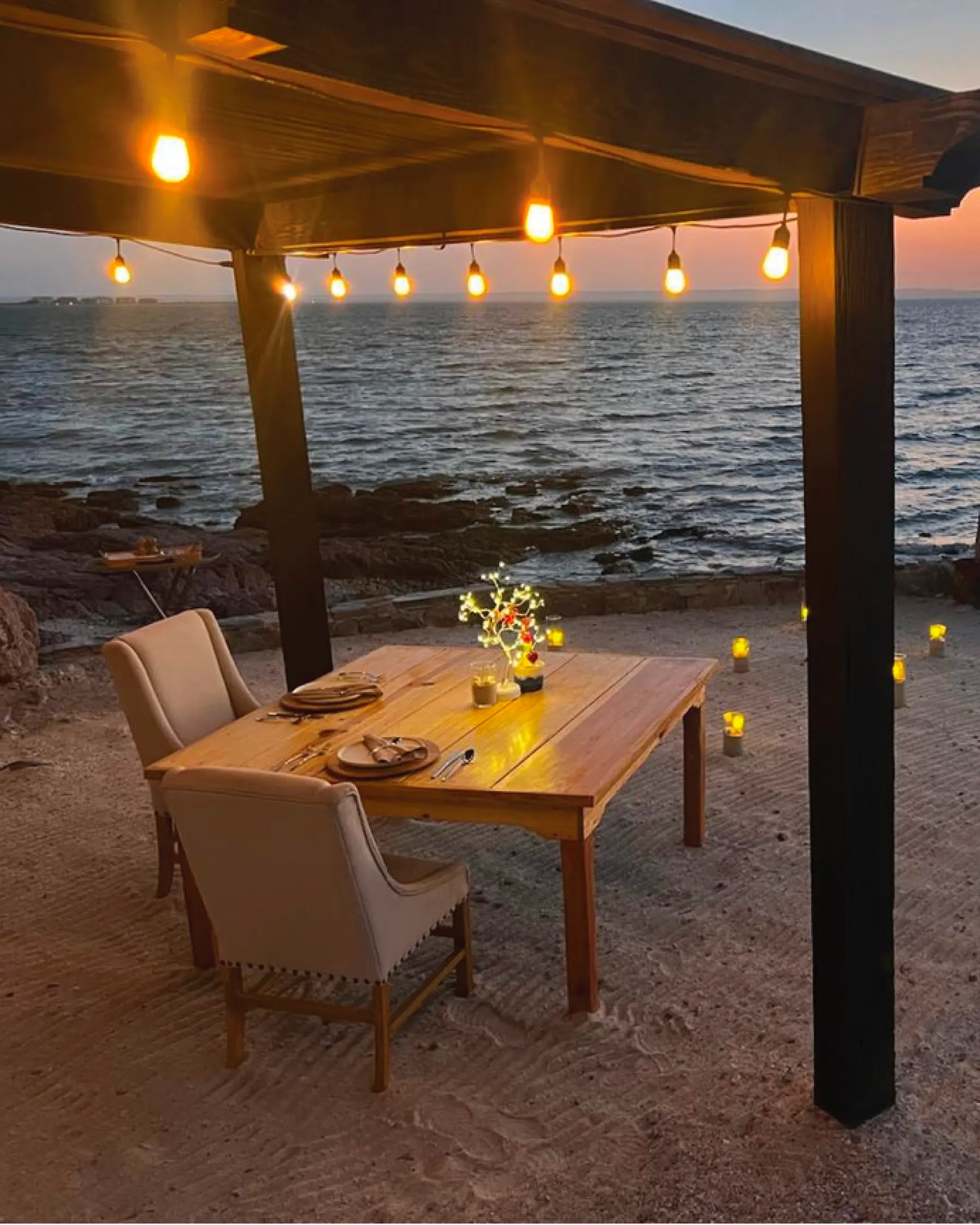 Wooden table set for two under a pergola strung with bulbs, on the sand facing the sea at dusk, with lit candle lanterns around it
