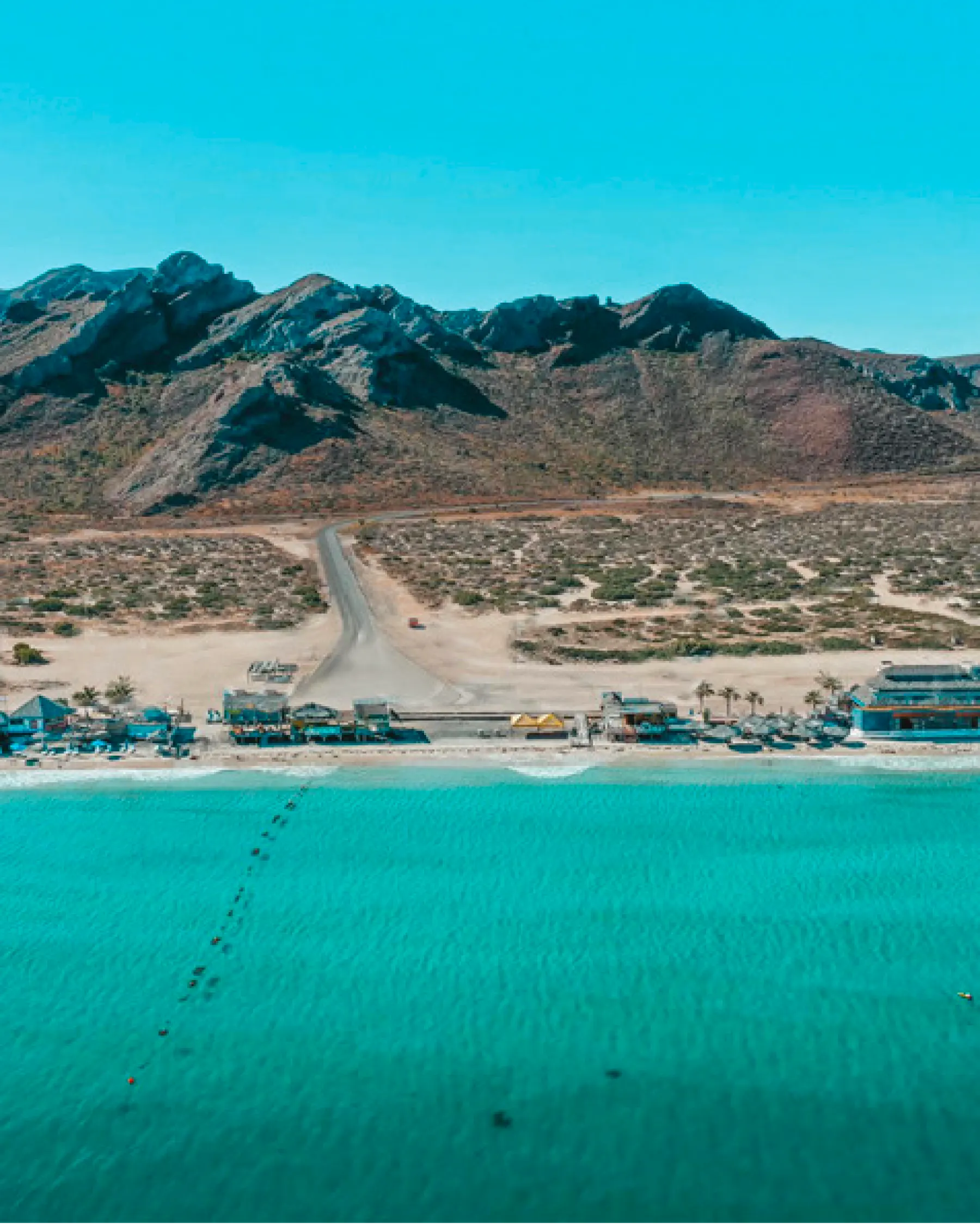 Aerial view of a turquoise-water beach with palapas and beach clubs along the shore, and desert hills behind