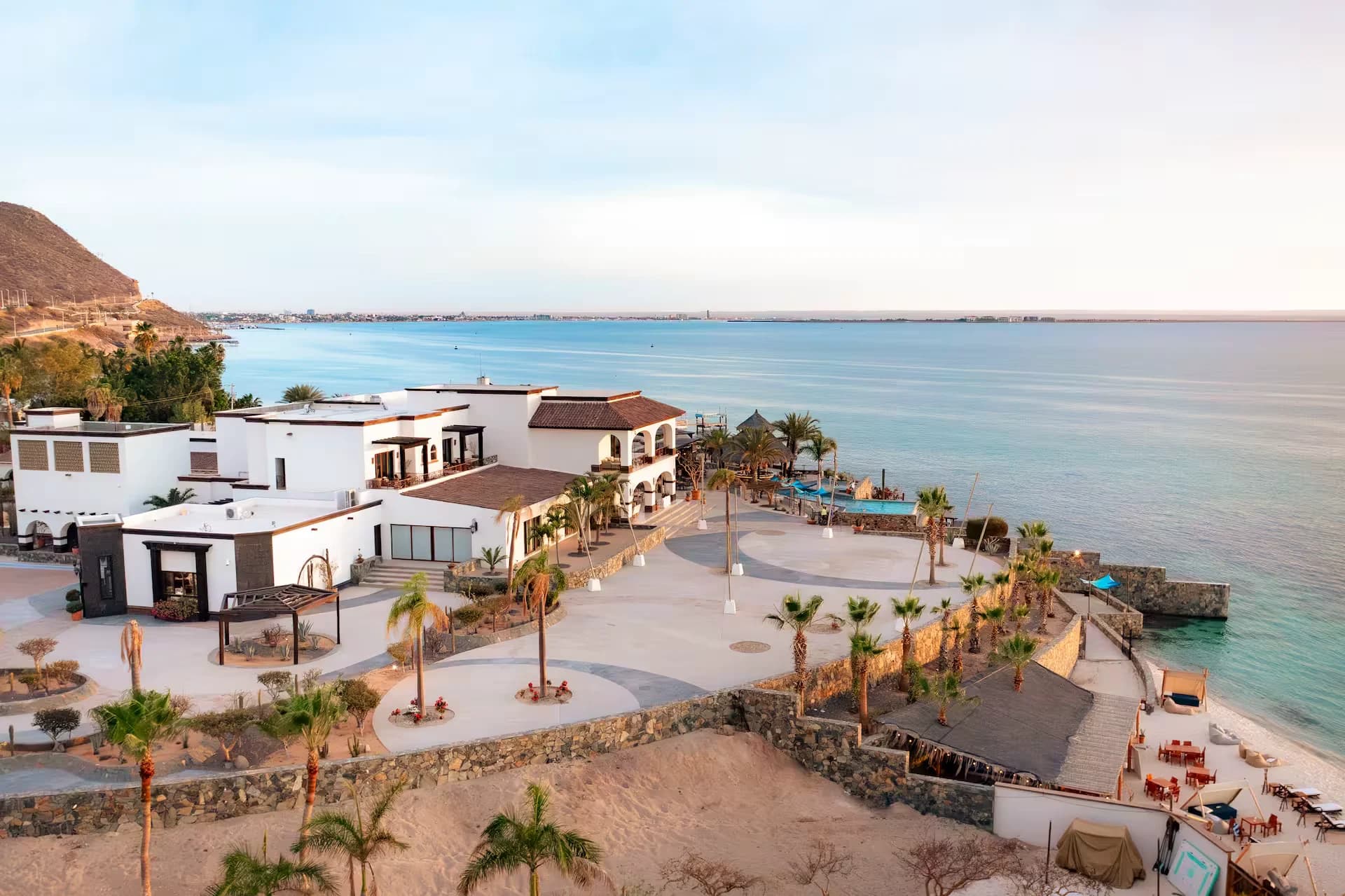 La casa vista desde arriba al atardecer: los techos de teja y las arcadas blancas, las palmeras y la alberca sobre la punta, con la bahía de La Paz y la ciudad al fondo