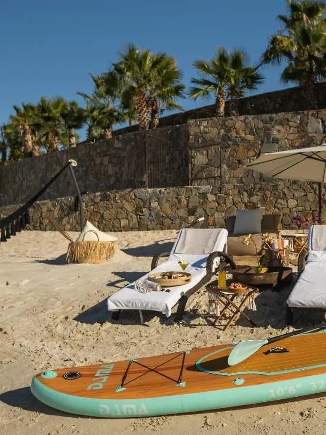 La playa de la casa con camastros, hamaca y sombrilla, una tabla de paddle en la arena y las palmeras del jardín detrás