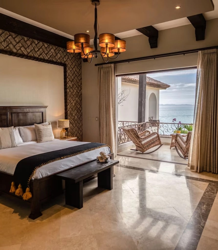 Dark timber king bed with a chandelier, and a balcony with rocking chairs beyond