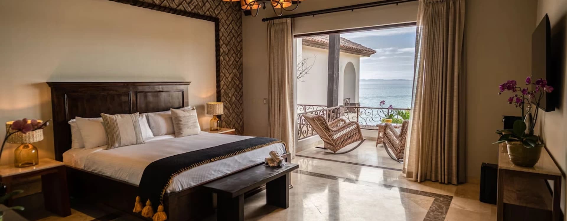 Dark timber king bed with a chandelier, and a balcony with rocking chairs beyond