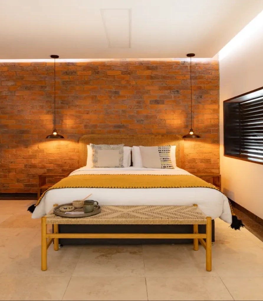 Bed with a bench at the foot under a brick wall and pendant lamps