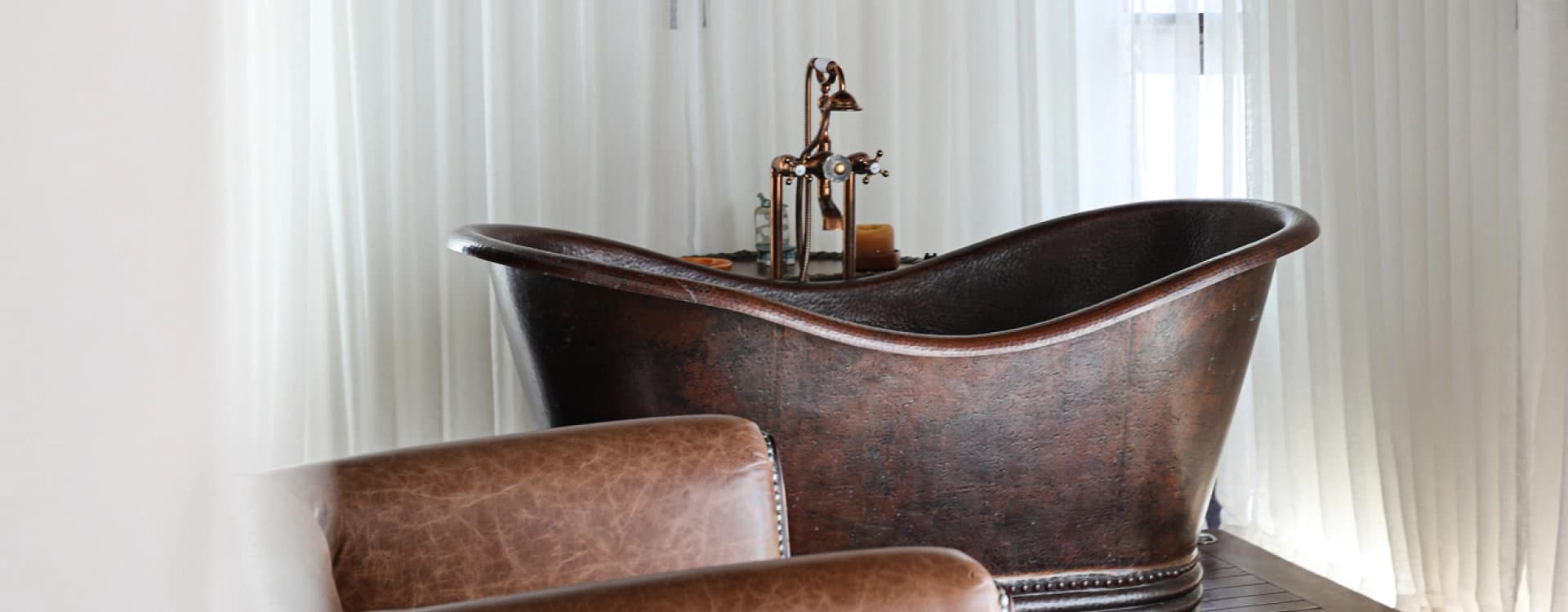 Freestanding copper tub before white curtains, with a leather armchair