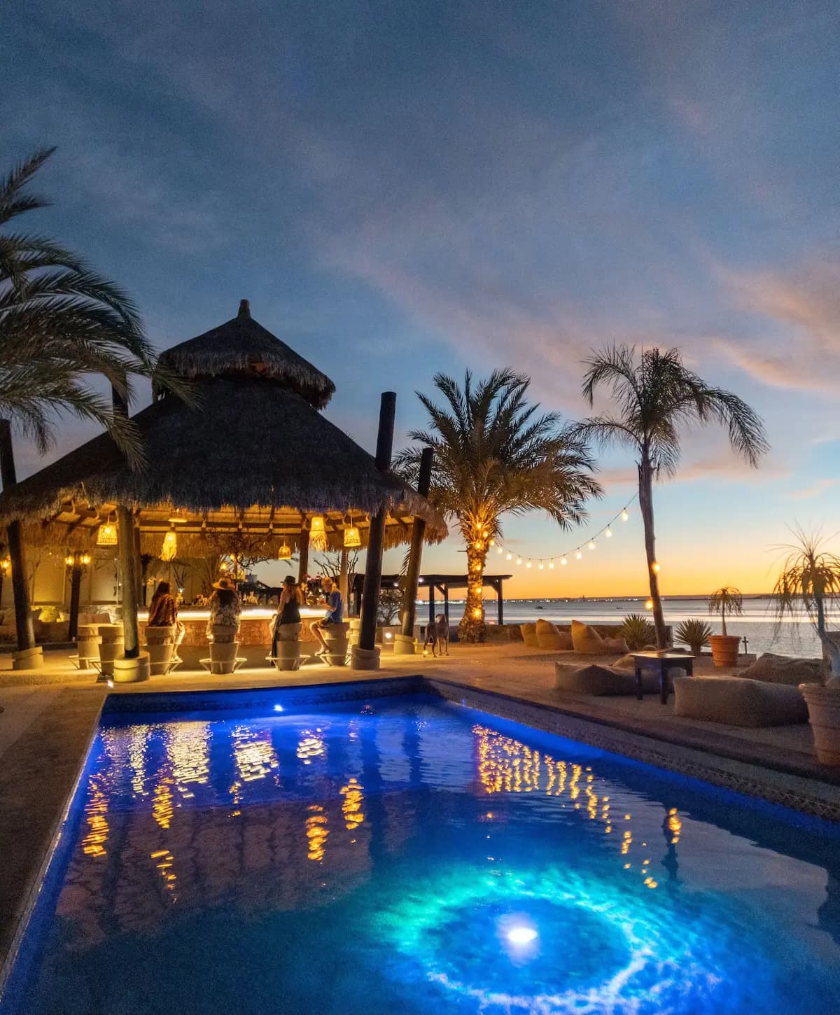 La barra de palapa al anochecer, con huéspedes sentados, luces de serie y la alberca frente al mar