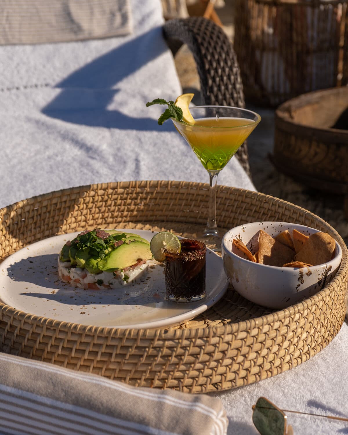 A wicker tray on a lounger, with an avocado ceviche, a cocktail and a small bowl of tortilla chips